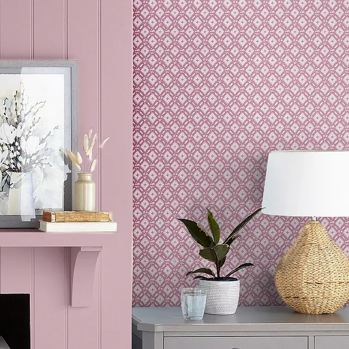 Laura Ashley Vliestapete Whitebrook - Rosa, Floral, 1.000 x 52 cm - Rosa Tapete mit wiederkehrendem weißem Geometermuster an einer Wand, dekoriert mit Lampe, Pflanze und Wohnaccessoires.