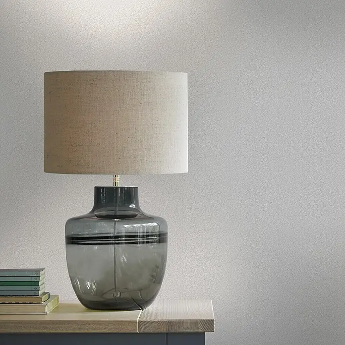 Laura Ashley Vliestapete Punktmuster - Weiß, Floral, 1.000 x 52 cm - Eine Tischlampe mit beigem Stoffschirm und rauchgrauem Glassockel steht auf einem Holztisch neben einem Stapel Bücher.