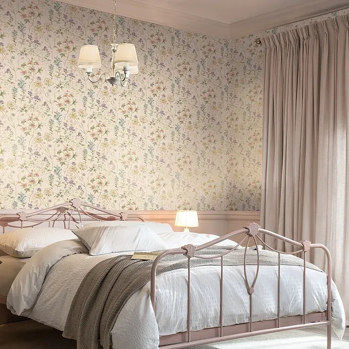 Laura Ashley Vliestapete Vliestapete Wildwiese - Rosa, Floral - Rosafarbenes Metallbettgestell mit weißer Bettwäsche in einem Schlafzimmer mit Blumenmuster-Tapete.