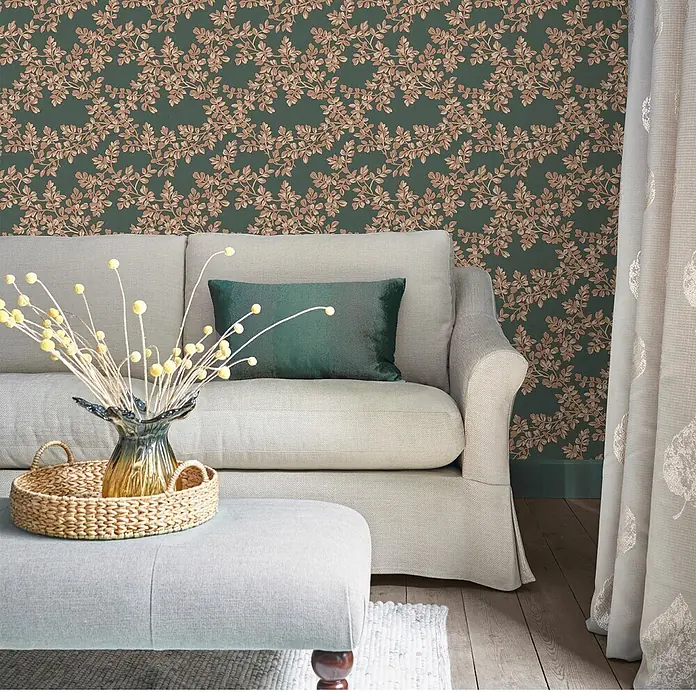 Laura Ashley Vliestapete Burnham - Dunkelgrün, Floral, 1.000 x 52 cm - Dunkelgrüne Tapete mit hellbraunem Blattmuster in einem Wohnzimmer, hinter einem hellen Sofa.
