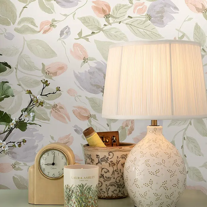 Laura Ashley Vliestapete Orisia-Pfingstrose - Grün, Floral, 1.000 x 52 cm - Weiße plissierte Lampe mit dekorativem Keramikfuß, die einen Tisch mit Uhr, Kerze und Blumendeko beleuchtet.