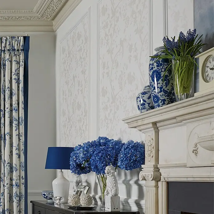Laura Ashley Vliestapete orientalischer Garten - Weiß, Floral, 1.000 x 52 cm - Ein klassisch eingerichtetes Zimmer mit blau-weißer Blumentapete, Vorhängen und Hortensien, mit einem verzierten Marmorkamin und Gesims.