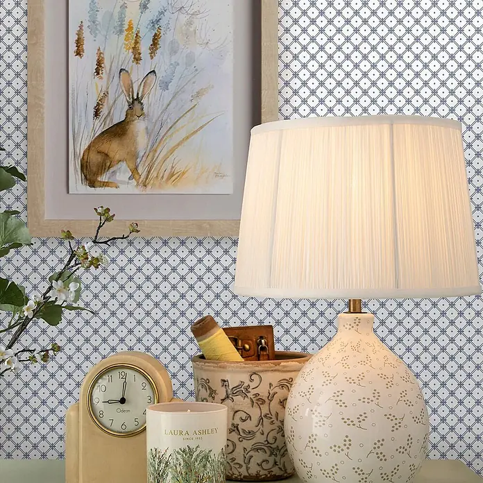 Laura Ashley Vliestapete Korbflechterei - Blau, Floral, 1.000 x 52 cm - Elfenbeinfarbene Keramiktischlampe mit goldenem Blumenmuster und plissiertem weißen Schirm, beleuchtet eine gemütliche Innenszene.