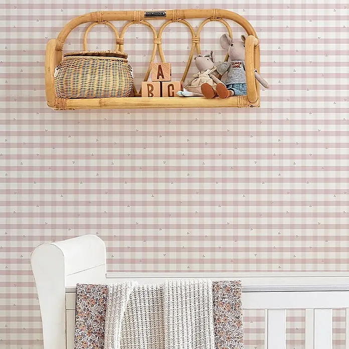 Laura Ashley Vliestapete Gingham - Rosa, Floral, 1.000 x 52 cm - Rosafarbene und weiße karierte Tapete im Kinderzimmer, mit Rattanregal und Spielzeug über einem weißen Kinderbett.