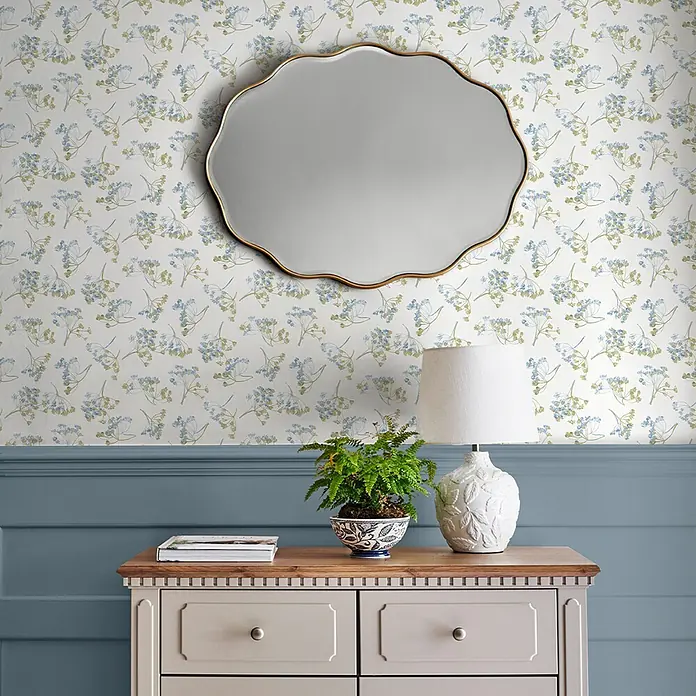 Laura Ashley Vliestapete Avonbury - Grün, Floral, 1.000 x 52 cm - Ein Raum mit weißer Blumentapete in Blau und Grün, einem goldgerahmten Wellenspiegel und einer beigen Kommode darunter.