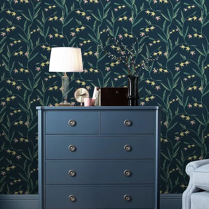 Laura Ashley Vliestapete Fuchsienblüten - Blau, Floral, 1.000 x 52 cm - Dunkelblaue botanische Tapete mit grünen Blättern und zarten gelb-rosa Blüten in einer Raumeinstellung.
