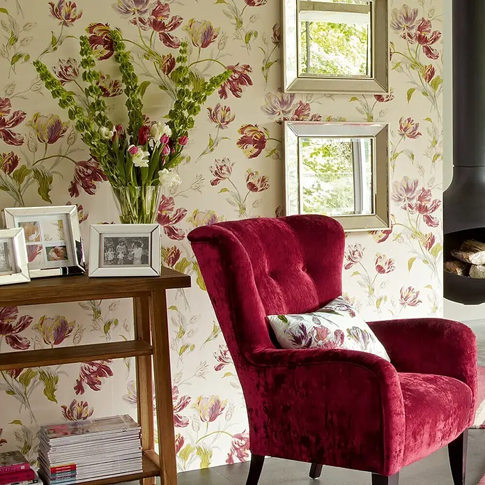 Laura Ashley Vliestapete Papageientulpen - Rot, Floral, 1.000 x 52 cm - Ein luxuriöser, tiefroter Samtsessel mit gemustertem Kissen in einem gemütlichen Raum.