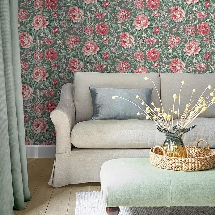 Laura Ashley Vliestapete Wildrosen - Grün, Floral, 1.000 x 52 cm - Florale Tapete mit rosa Rosen und grünen Blättern auf dunkelgrünem Grund, ziert eine Wohnzimmerwand hinter einem beigen Sofa.