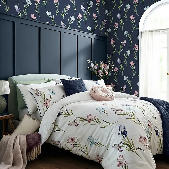 Laura Ashley Vliestapete Irisblüten - Blau, Floral, 1.000 x 52 cm - Florale Bettwäschegarnitur mit Aquarell-Iris in Pink, Blau und Lila auf weißem Grund, auf einem Bett dekoriert.
