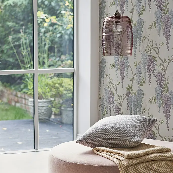 Laura Ashley Vliestapete Glyziniengarten - Blau/Rosa, Floral, 1.000 x 52 cm - Ein runder rosa Pouf mit einem grauen Kissen und einer gelben Decke darauf, neben einem Fenster.