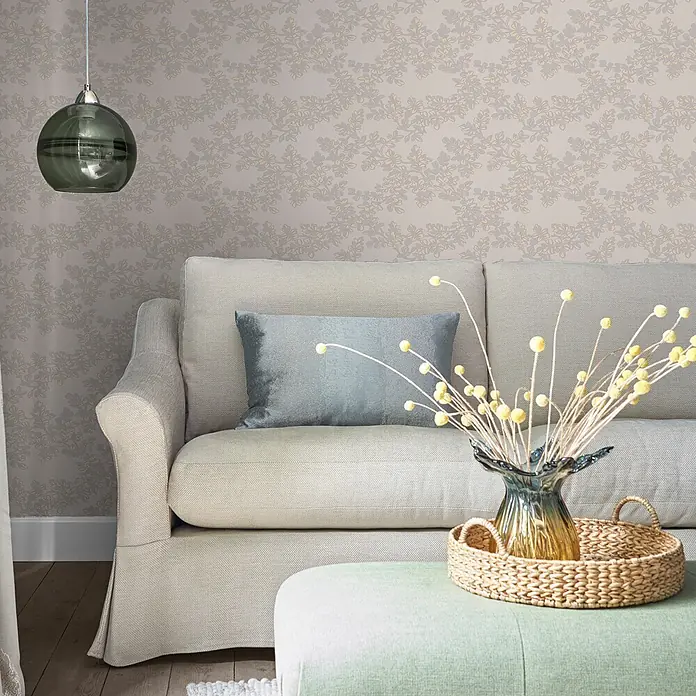 Laura Ashley Vliestapete Burnham - Hellgrau, Floral, 1.000 x 52 cm - Gemütliches Wohnzimmer mit hellbeigem Sofa, blauem Kissen, hellgrünem Hocker mit Korbtablett, gelben Trockenblumen und floraler Tapete.
