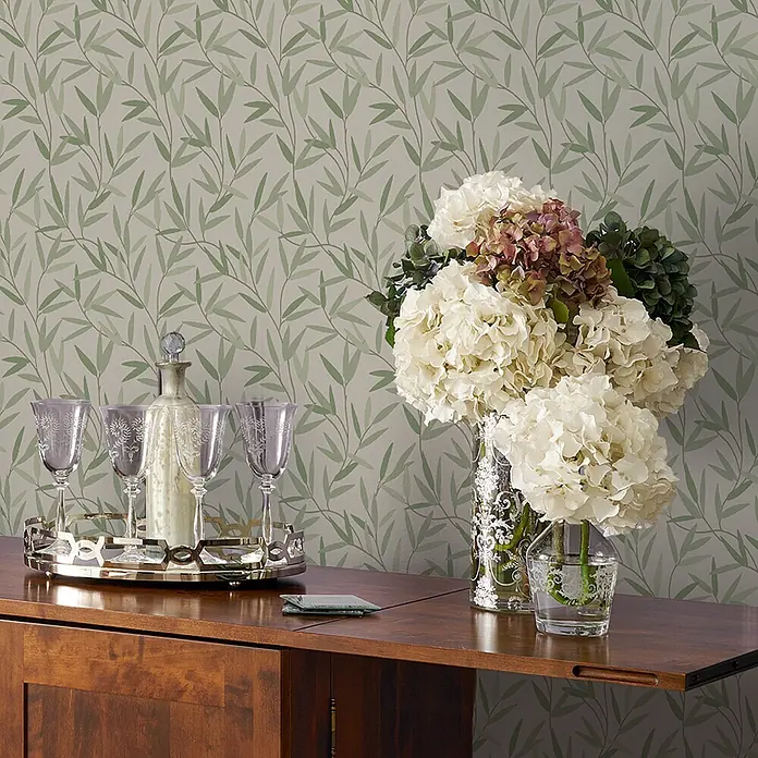 Laura Ashley Vliestapete Weidenblatt - Grün, Floral, 1.000 x 52 cm - Tapete mit einem sich wiederholenden Muster aus grünen Bambusblättern, schmückt eine Wand hinter einem Holzschrank mit Blumen.