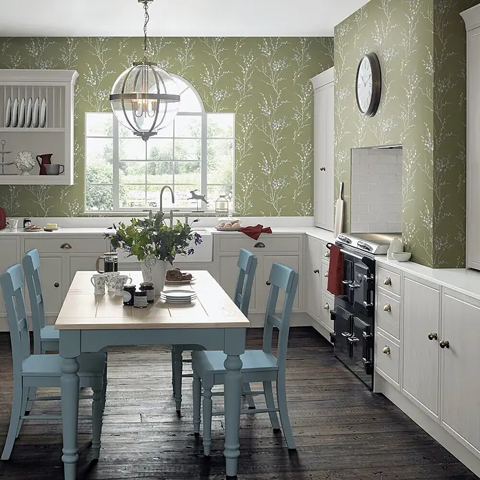 Laura Ashley Vliestapete Weidenkätzchen - Grün, Floral, 1.000 x 52 cm - Eine helle Küche mit grüner Blumenmuster-Tapete, hellen Cremeschränken, einem hellblauen Esstisch mit Stühlen und dunklem Holzboden.