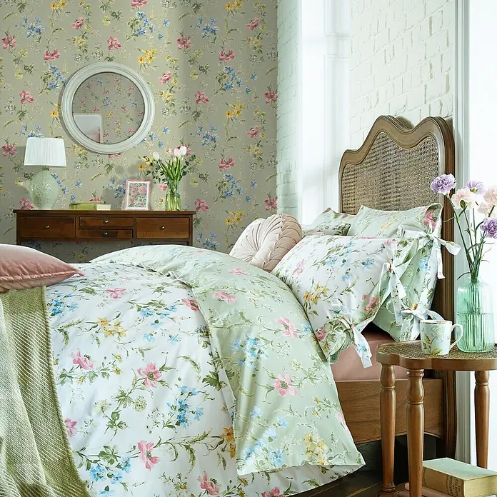 Laura Ashley Vliestapete Mohnwiese - Grün, Ornament, 1.000 x 52 cm - Florale Bettwäschegarnitur mit rosa, blauen und gelben Blumen auf hellgrünem Bett in gemütlichem Schlafzimmer.