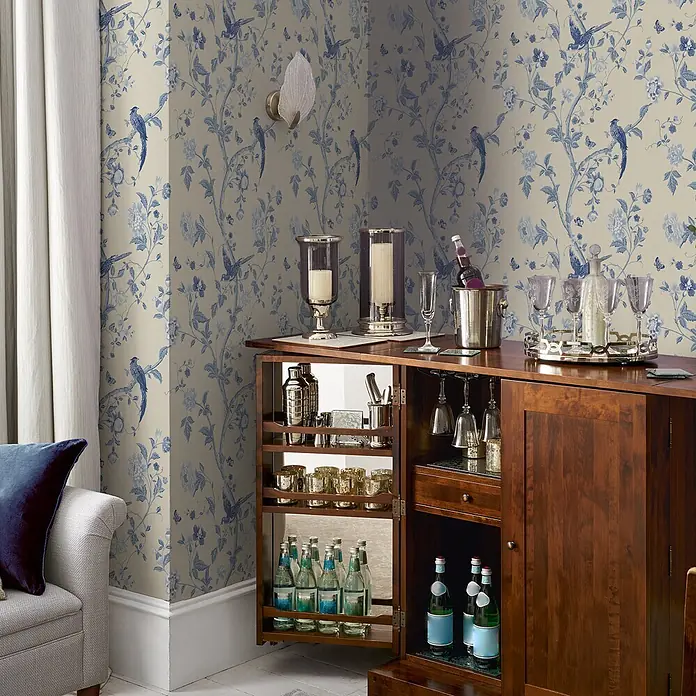 Laura Ashley Vliestapete Sommerpalast - Blau, Floral, 1.000 x 52 cm - Dunkler Holzbarschrank mit Getränken, Gläsern und Barzubehör, vor blau-beiger Tapete in einem Raum.
