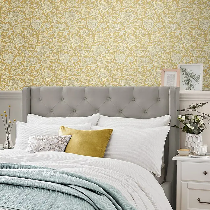 Laura Ashley Vliestapete Vliestapete Trailing Laurissa - Gelb, Floral - Ein gemütliches Schlafzimmer mit gelber Blumentapete, einem grauen gepolsterten Kopfteil und einem Bett mit weißer und blauer Steppbettwäsche.