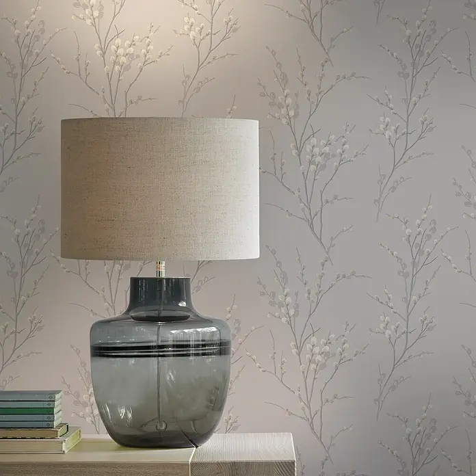 Laura Ashley Vliestapete Weidenkätzchen - Hellgrau, Floral, 1.000 x 52 cm - Tischleuchte mit natürlichem Stoffschirm und grauem Glassockel, die einen Nachttisch mit Büchern beleuchtet.
