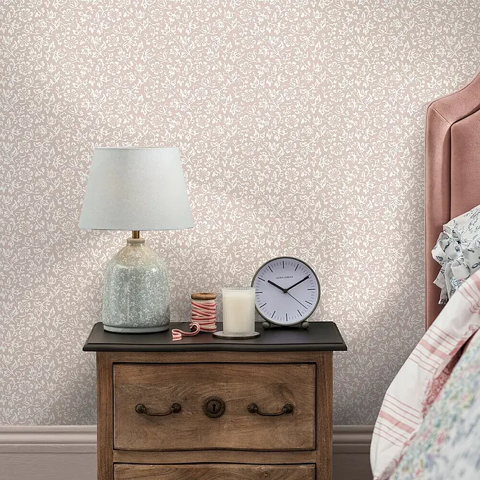 Laura Ashley Vliestapete Süßes Alyssum - Rosa, Floral, 1.000 x 52 cm - Beigefarbene Tapete mit weißem Blumenmuster an einer Schlafzimmerwand hinter einem Nachttisch.
