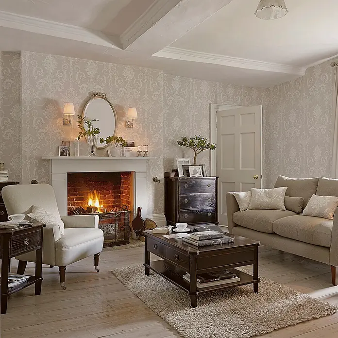 Laura Ashley Vliestapete Vliestapete Josette Damast - Hellgrau/Weiß, Ornament - Ein gemütliches Wohnzimmer mit hell gemusterter Tapete, einem beigen Sofa und Sessel und dunklen Holzmöbeln um einen beleuchteten Kamin.