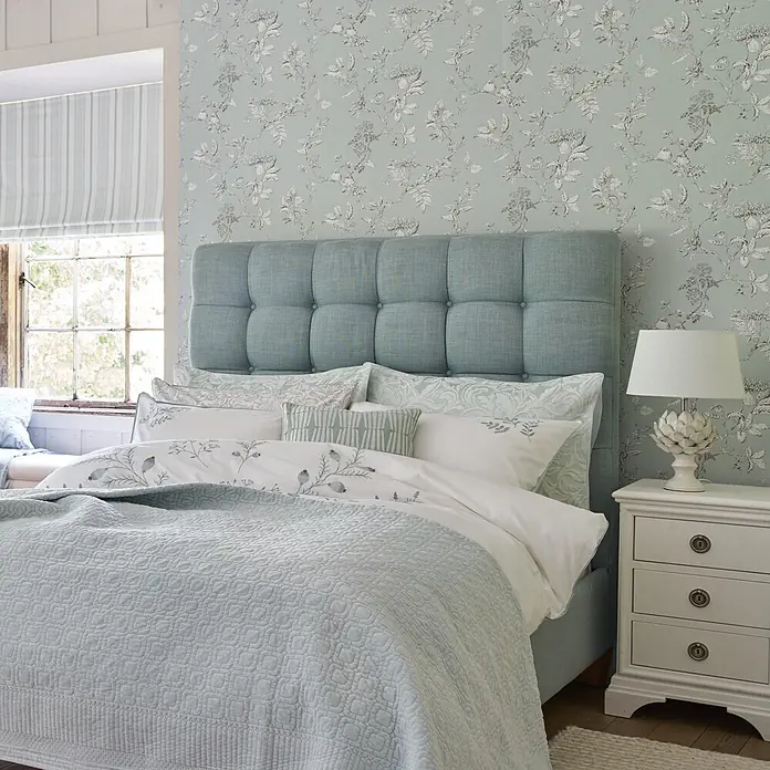 Laura Ashley Vliestapete Vliestapete Elderwood - Blau, Floral, 10 x 0,52 m - Ein ruhiges Schlafzimmer mit hellblauem getuftetem Kopfteil, weiß-blau gemusterter Bettwäsche und hellblauer Steppdecke.
