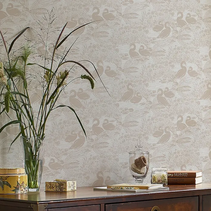 Laura Ashley Vliestapete Swans - Grau, Tieroptik, 1.000 x 52 cm - Beigefarbene Tapete mit Schwänen und Seerosenmuster an einer Wand hinter einer Kommode mit Pflanzen und Dekoration.