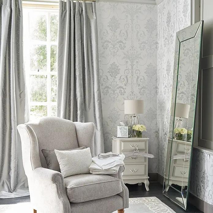 Laura Ashley Vliestapete Josette Damast - Silber, Ornament, 1.000 x 52 cm - Ein ruhiger Wohnraum mit grauem Sessel und Kissen, schimmernden grauen Vorhängen, einem Ganzkörperspiegel und einem weißen Nachttisch mit Lampe.