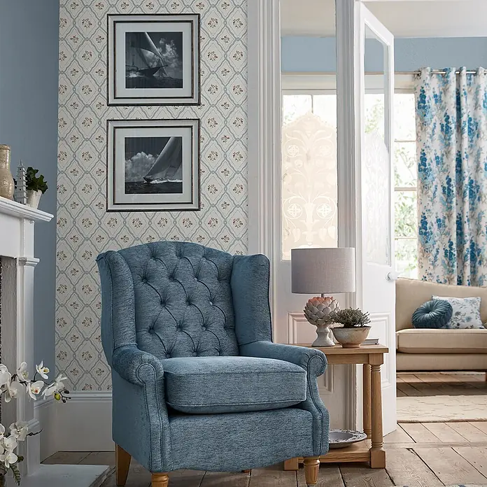 Laura Ashley Vliestapete Pinford Spalier - Hellblau, Floral, 1.000 x 52 cm - Ein blauer Sessel mit Knopfheftung aus strukturiertem Stoff in einem gemütlichen Wohnzimmer.