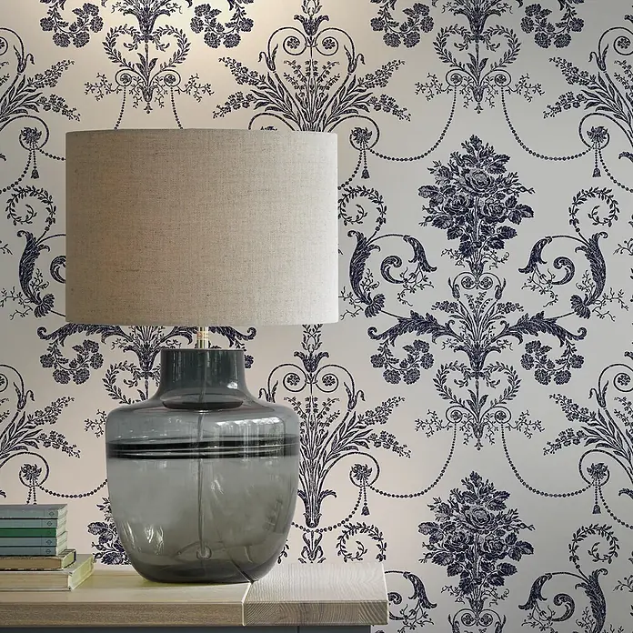 Laura Ashley Vliestapete Vliestapete Josette Damast - Dunkelblau/Weiß, Ornament, 10 x 0,52 m - Graue Glas-Tischlampe mit beigem Stoffschirm, steht auf einem hellen Holztisch vor einer gemusterten Tapete.