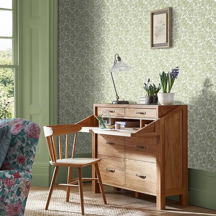 Laura Ashley Vliestapete blühende Hortensie - Grün, Floral, 1.000 x 52 cm - Sekretär aus Naturholz mit ausziehbarer Schreibfläche und passendem Stuhl in einem hellen, gemusterten Raum.