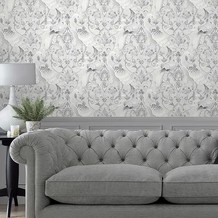 Laura Ashley Vliestapete Tregaron - Silber, Ornament, 1.000 x 52 cm - Grau getuftetes Stoffsofa in einem Wohnzimmer mit weiß-grauer Damast-Tapete mit Pfauenmotiv.