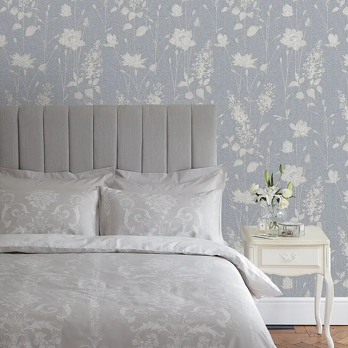 Laura Ashley Vliestapete Vliestapete Libellen Garten - Blau, Floral - Schlafzimmer mit Bett, grauem gepolstertem Fischgrät-Kopfteil und hellgrauer gemusterter Bettwäsche mit Kissen.