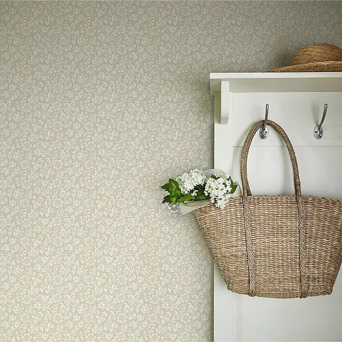 Laura Ashley Vliestapete Süßes Alyssum - Naturbelassen, Floral, 1.000 x 52 cm - Hellbeigefarbene Tapete mit zartem weißem Blumenmuster, die eine Wand neben einer weißen Garderobe mit Strohtasche und Hut schmückt.