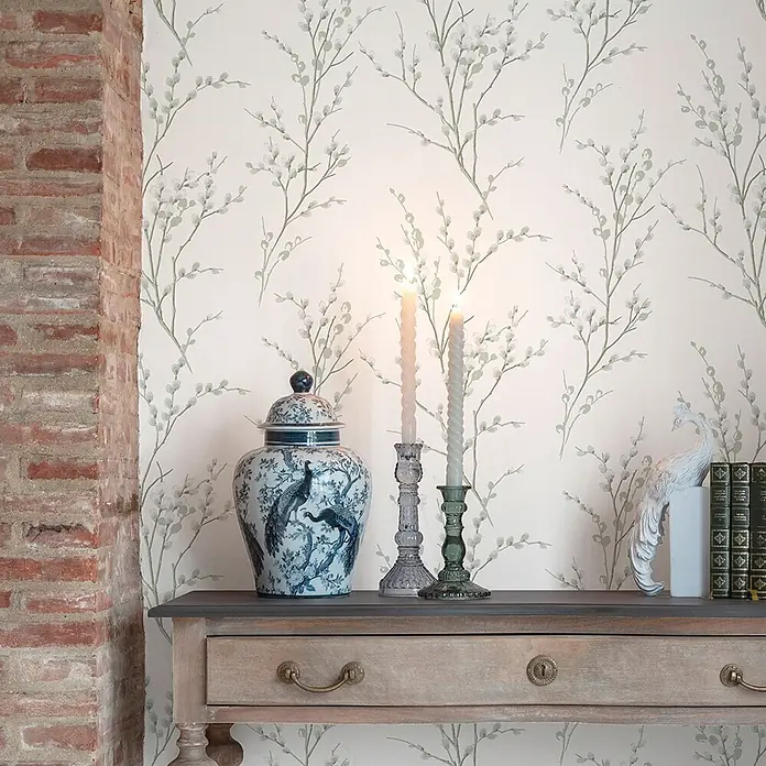 Laura Ashley Vliestapete Weidenkätzchen - Grün, Floral, 1.000 x 52 cm - Cremefarbene Tapete mit zartem grünem Weidenkätzchenmuster an einer Wand neben einem Ziegelpfeiler und Konsolentisch.