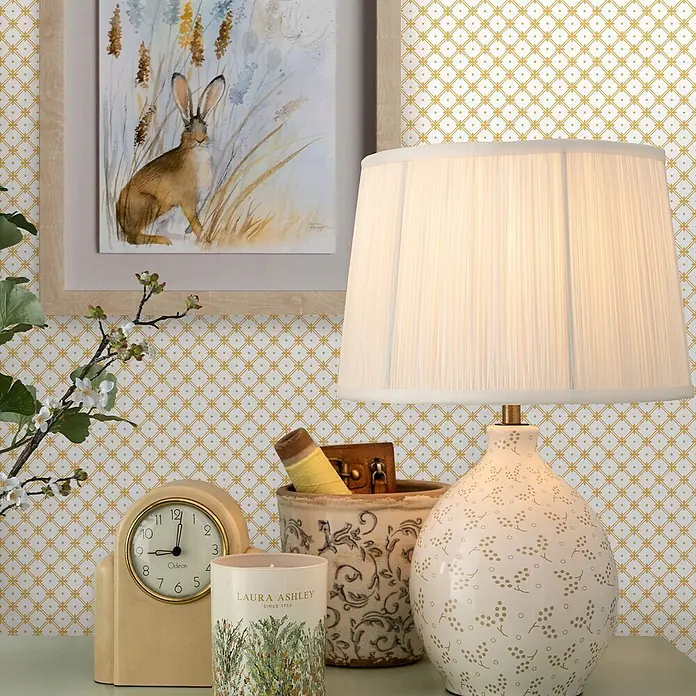 Laura Ashley Vliestapete Flechtwerk - Gelb/Ocker, Ornament, 1.000 x 52 cm - Ein Zimmer mit weißer Tapete mit goldenem geometrischem Muster, einem gerahmten Hasenbild und einer plissierten Lampe.