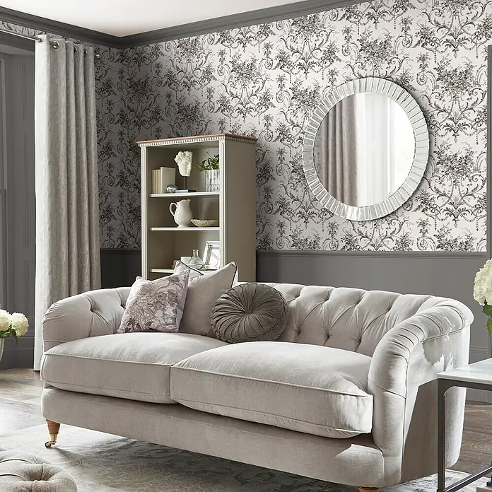 Laura Ashley Vliestapete Tuileries Französisch - Grau, Floral, 1.000 x 52 cm - Ein hellbeiges getuftetes Sofa mit Plüschkissen in einem stilvollen Wohnzimmer.