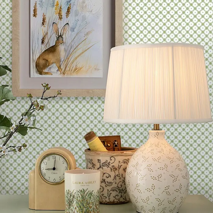 Laura Ashley Vliestapete Vliestapete Korbflechterei - Grün, Ornament, 1.000 x 52 cm - Eine weiße Keramiktischlampe mit gemustertem Sockel und plissiertem Schirm, beleuchtet auf einem Beistelltisch.