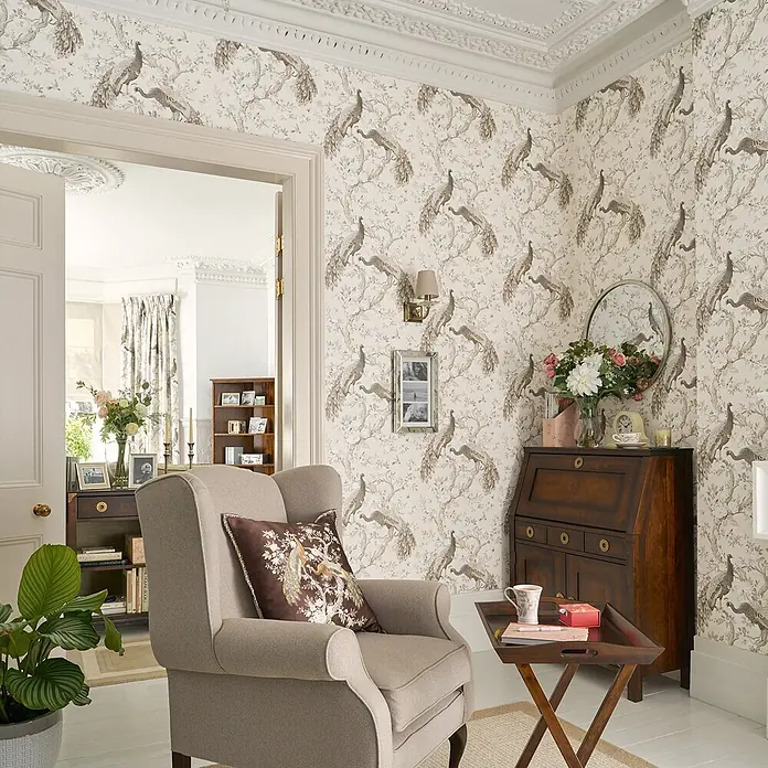 Laura Ashley Vliestapete Belvedere Pfau - Braun, Floral, 1.000 x 52 cm - Cremefarbene Tapete mit einem detaillierten Muster aus braunen Pfauen und blühenden Bäumen, die Wände eines klassischen Wohnzimmers schmückt.