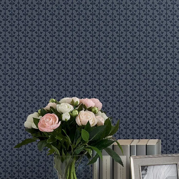 Laura Ashley Vliestapete Seaham - Blau, Grafisch, 1.000 x 52 cm - Dunkelblaue Tapete mit subtilem, wiederholtem Rankenmuster aus Herzen, gezeigt an einer Wand hinter Blumen und Büchern.