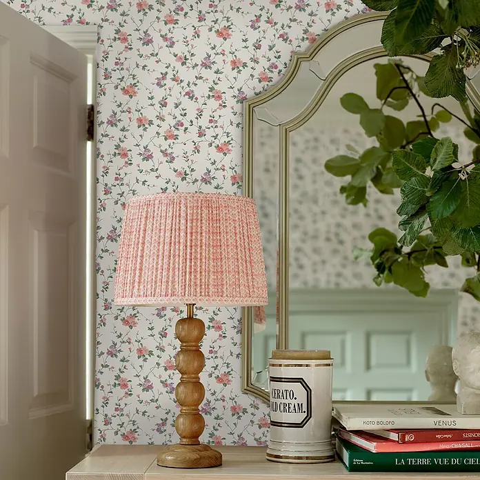 Laura Ashley Vliestapete Vliestapete Priorat - Rosa, Floral, 10 x 0,52 m - Eine Holz-Tischlampe mit plissiertem rosa gemustertem Lampenschirm auf einem hellen Holztisch.