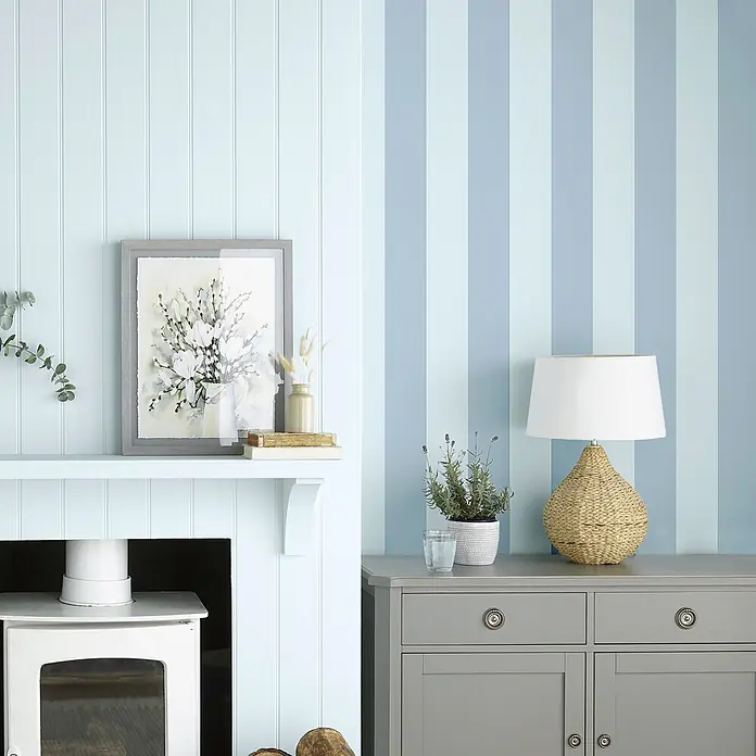 Laura Ashley Vliestapete Lille Streifen - Blau, Streifen, 1.000 x 52 cm - Ein gemütlicher Raum mit hellblauen Vertäfelungen und blau gestreifter Tapete, eingerichtet mit einem grauen Schrank und einem weißen Ofen.