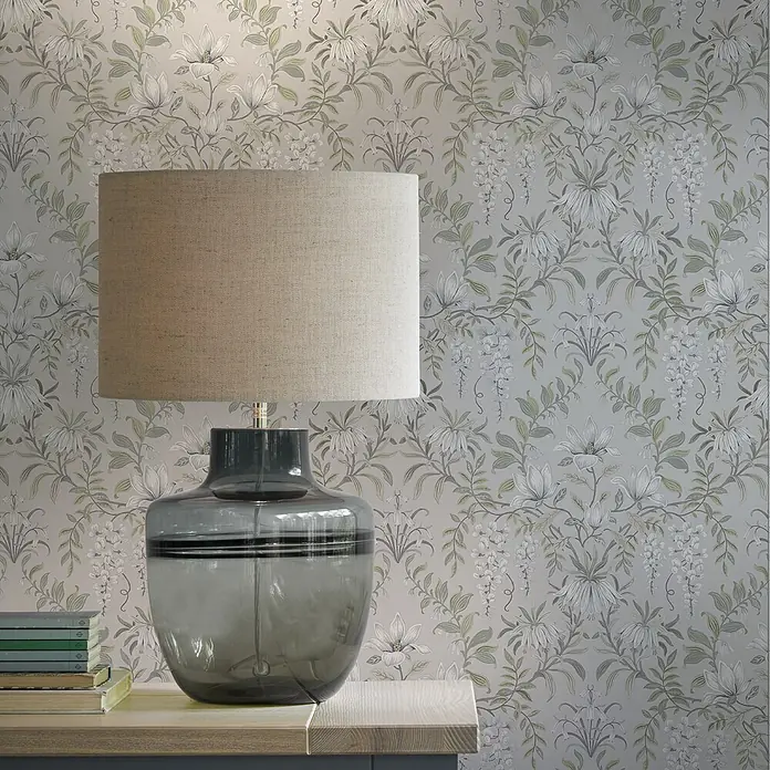 Laura Ashley Vliestapete Vintage Blumen - Salbeigrün, Floral, 1.000 x 52 cm - Tischlampe mit dunkelgrauem Glasfuß und beigem Stoffschirm, auf einer hellen Holzoberfläche stehend.