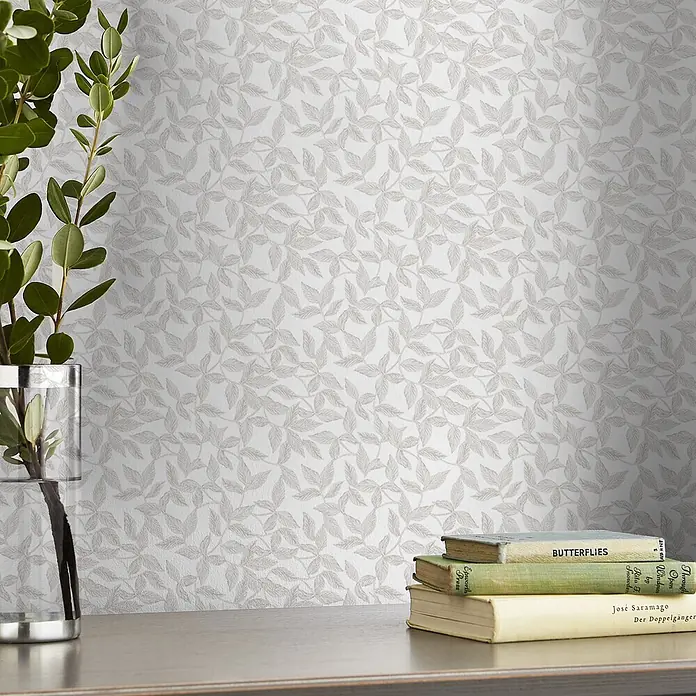 Laura Ashley Vliestapete Erwood - Grau, Floral, 1.000 x 52 cm - Weiße Tapete mit hellem, geprägtem Blattmuster, in einem Raum mit Pflanze und Büchern.
