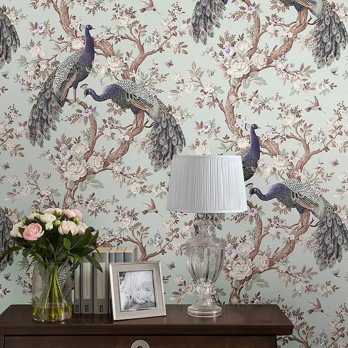Laura Ashley Vliestapete Belvedere Pfau - Blau, Tieroptik, 1.000 x 52 cm - Tapete mit Chinoiserie-Muster, das kunstvolle Pfauen, blühende Bäume, fliegende Vögel und Schmetterlinge auf hellgrünem Hintergrund zeigt, an einer Wand.