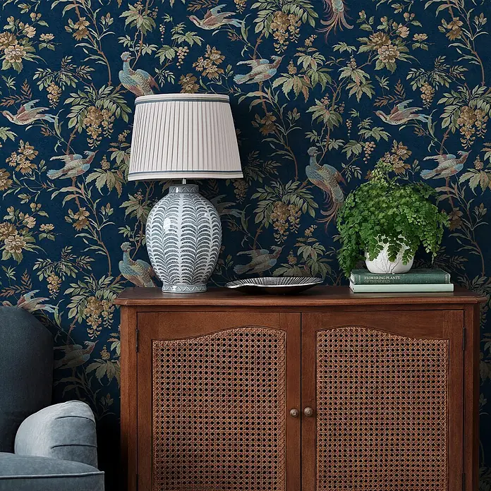 Laura Ashley Vliestapete Attleton Blume - Blau, Floral, 1.000 x 52 cm - Dunkelblaue Tapete mit aufwendigen Chinoiserie-Vogel- und Blumenmotiven an einer Wand hinter Möbeln.