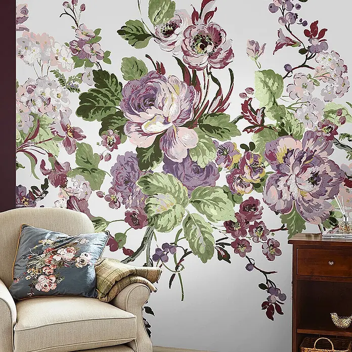 Laura Ashley Fototapete - Vlies - Großes florales Wandbild mit lila, rosa, grünen und weißen Blumen, das sich über eine Zimmerwand über einem Sessel und Beistelltisch erstreckt.