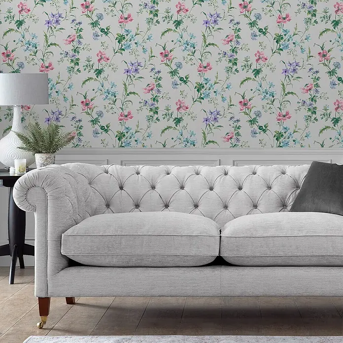 Laura Ashley Vliestapete Mohnwiese - Lila, Floral, 1.000 x 52 cm - Hellgraues Chesterfield-Sofa mit gerollten Armlehnen, Fischgrätstoff, Holzbeinen und Messingrollen, in einem Wohnzimmer.
