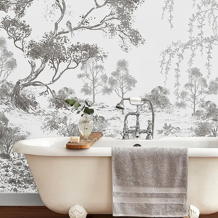 Laura Ashley Fototapete - Vlies - Eine klassische weiße freistehende Badewanne mit grauem Handtuch, einem Holztablett mit Seife und Pflanze, vor einer Tapete mit Naturmotiv.