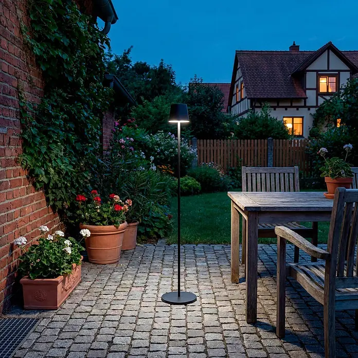 Starlux Paola Akku-Stehleuchte - Schwarz, RGBW, 3 W, IP44 - Schwarze Outdoor-Stehlampe beleuchtet eine gepflasterte Terrasse in der Dämmerung, umgeben von Pflanzen und einem Haus.