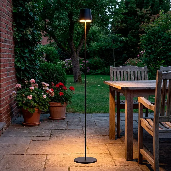 Starlux Paola Akku-Stehleuchte - Schwarz, RGBW, 3 W, IP44 - Schwarze Outdoor-Stehleuchte auf Steinplatte, die in einem Garten warmes Licht auf den Boden wirft.