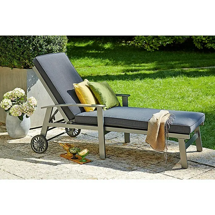Sunfun Maja Gartenliege - B x T x H: 170,5 x 63,5 x 102 cm, Aluminium, Anthrazit, Rollbar - Ein grauer Liegestuhl mit Rädern, Kissen und einer Decke steht auf einer Terrasse neben Blumen und Grün.
